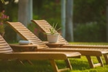 Three wooden lounge chairs are arranged on a grassy lawn, with sunlight casting shadows on them. A small table between the chairs holds a white potted plant, and there is a small white dish on one of the chairs.