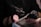 A person wearing black gloves is holding a rotary tool while working on someone's nails. The setting suggests a nail salon or a nail art process. The person receiving the service has their hand resting on a padded surface, and a pink makeup brush is visible in the scene.