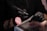 A person wearing black gloves is holding a rotary tool while working on someone's nails. The setting suggests a nail salon or a nail art process. The person receiving the service has their hand resting on a padded surface, and a pink makeup brush is visible in the scene.