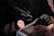 A person wearing black gloves is holding a rotary tool while working on someone's nails. The setting suggests a nail salon or a nail art process. The person receiving the service has their hand resting on a padded surface, and a pink makeup brush is visible in the scene.