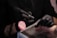 A person wearing black gloves is holding a rotary tool while working on someone's nails. The setting suggests a nail salon or a nail art process. The person receiving the service has their hand resting on a padded surface, and a pink makeup brush is visible in the scene.