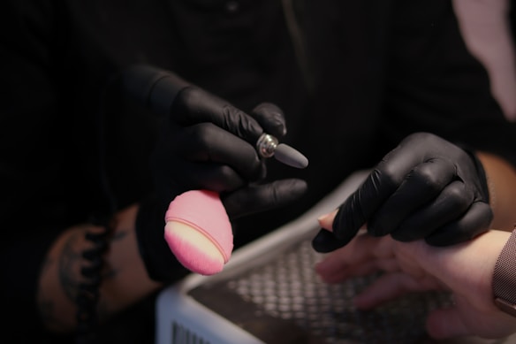 A person wearing black gloves is holding a rotary tool while working on someone's nails. The setting suggests a nail salon or a nail art process. The person receiving the service has their hand resting on a padded surface, and a pink makeup brush is visible in the scene.