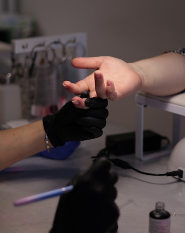 Two hands are visible, with one wearing black gloves holding the fingers of the other hand, suggesting a manicure or nail care session. The background includes various manicure tools and equipment. The setting appears to be a nail salon or spa environment.