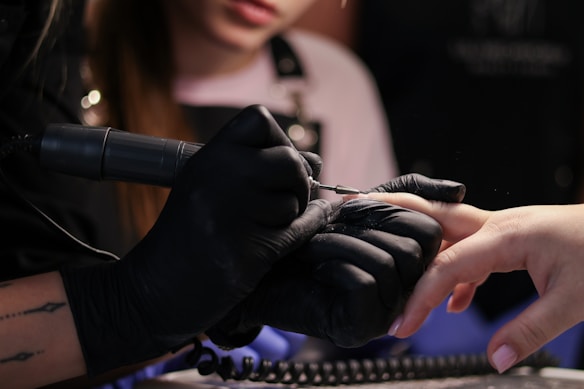 A person wearing black gloves is using a nail drill on someone else's finger. The focus is on the hands and the nail treatment process, suggesting a manicure or nail care session.