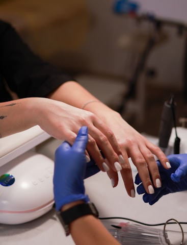 A close-up view of hands receiving a manicure, with a technician wearing blue gloves applying nail care. The nails are polished and well-groomed.