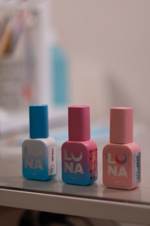 Three bottles of nail polish are displayed in a row on a reflective surface. The bottles are cylindrical with square bases, each with a distinct color theme. The one on the left is predominantly blue, the middle one is pink, and the right one is a lighter pink. All bottles feature the brand name 'LUNA' in large, bold letters.