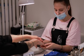 A person wearing a face mask and an apron is focused on performing a manicure for a client whose hands are extended towards them. The setting appears to be a nail salon, with a lamp providing light and manicure tools visible on the table.