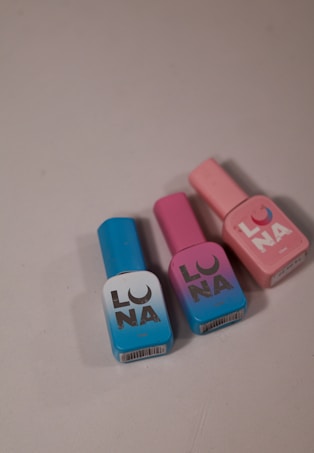 Three bottles of nail polish are arranged on a light-colored surface. The bottles are in blue, purple, and pink colors and each one has 'LUNA' written on the front in a bold font.