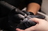 A close-up of a nail technician wearing black gloves using an electric nail file on a client's fingernail. Dust from the filing process is visible around the tool in use.