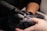 A close-up of a nail technician wearing black gloves using an electric nail file on a client's fingernail. Dust from the filing process is visible around the tool in use.