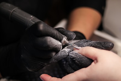 A close-up of a nail technician wearing black gloves using an electric nail file on a client's fingernail. Dust from the filing process is visible around the tool in use.