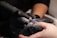 A close-up of a nail technician wearing black gloves using an electric nail file on a client's fingernail. Dust from the filing process is visible around the tool in use.