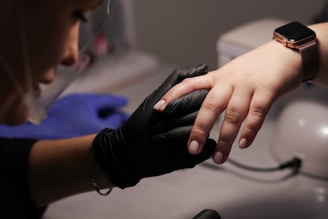A person wearing black gloves holds another person's hand, focusing on the fingernails. The person receiving the hand treatment wears a smartwatch with a metal band.