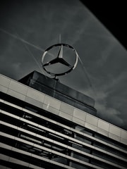 A large metallic logo in the shape of a three-pointed star is mounted on top of a modern building. The structure of the building features horizontal lines of windows and metal panels, reflecting the subdued light. The sky in the background is overcast, contributing to a somber and industrial atmosphere.