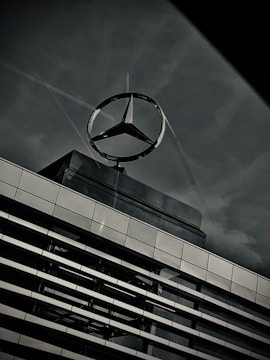 A large metallic logo in the shape of a three-pointed star is mounted on top of a modern building. The structure of the building features horizontal lines of windows and metal panels, reflecting the subdued light. The sky in the background is overcast, contributing to a somber and industrial atmosphere.