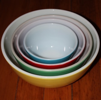 A set of ceramic bowls is nested together, each a different color, from yellow to white, to create a pleasing gradient. The bowls are placed on a wooden surface.