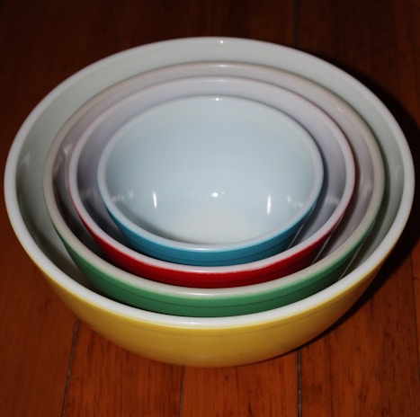 A set of ceramic bowls is nested together, each a different color, from yellow to white, to create a pleasing gradient. The bowls are placed on a wooden surface.
