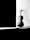 A violin is placed upright on the floor against a light-colored, possibly white, wall next to a dark, almost black, rectangular object or shadow. The image is in black and white, creating a stark contrast between the lit and shadowed areas.