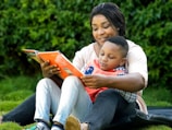 An adult and a child sit together on a grassy area, deeply engaged in reading a book. The adult, who is smiling warmly, holds the book and embraces the child, who appears focused on the pages. Both wear casual clothing, and their expressions reflect a shared moment of learning and connection.