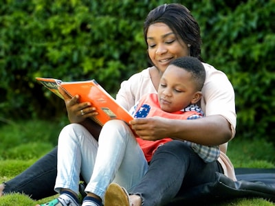 An adult and a child sit together on a grassy area, deeply engaged in reading a book. The adult, who is smiling warmly, holds the book and embraces the child, who appears focused on the pages. Both wear casual clothing, and their expressions reflect a shared moment of learning and connection.