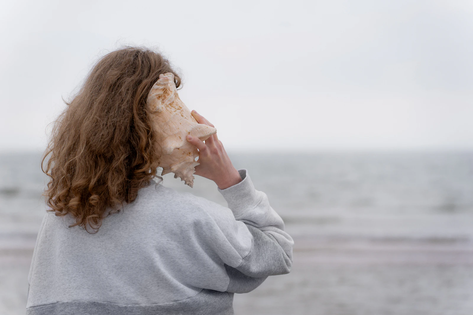 Frau hält eine Muschel ans Ohr und blickt aufs Meer