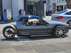 A unique, sporty three-wheeled vehicle with sleek design and large rims is parked on a city street. Pedestrians are visible in the background, and a modern building with storefronts can be seen.