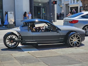 A unique, sporty three-wheeled vehicle with sleek design and large rims is parked on a city street. Pedestrians are visible in the background, and a modern building with storefronts can be seen.
