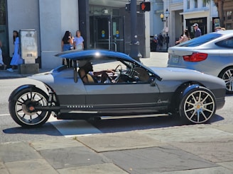 A unique, sporty three-wheeled vehicle with sleek design and large rims is parked on a city street. Pedestrians are visible in the background, and a modern building with storefronts can be seen.