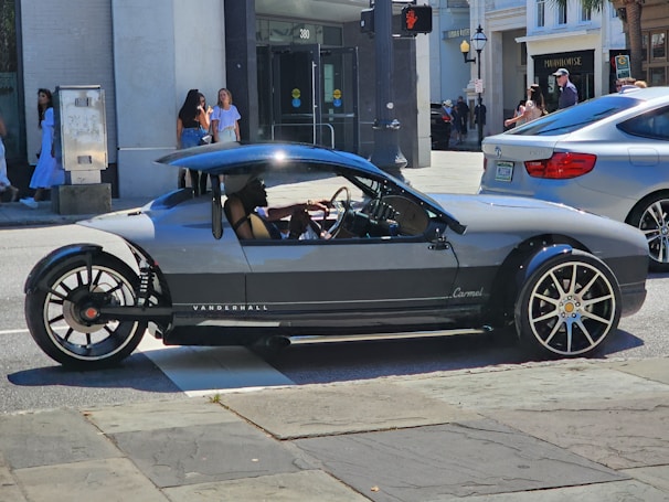 A unique, sporty three-wheeled vehicle with sleek design and large rims is parked on a city street. Pedestrians are visible in the background, and a modern building with storefronts can be seen.