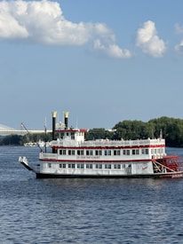A paddleboat with multiple decks and red, white, and black colors is sailing on a calm body of water. The boat has two large yellow chimneys and a traditional paddle wheel at the back. In the background, there is a green treeline and a bridge partially visible on the left side. The sky is clear with a few white, fluffy clouds.