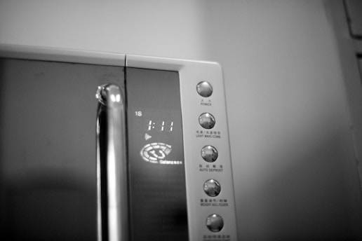 A microwave oven control panel features a digital display showing the time 1:11. Several buttons for different functions are arranged vertically, with labels in both Chinese and English. The microwave's sleek design includes a metallic handle, reflecting the light faintly.