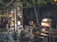 A market stall located outdoors under the shade of trees, displaying various electrical appliances and goods. The stand includes items like fans and lamps, with some being illuminated. The vendor, an elderly individual wearing glasses, sits amidst the array of products. The scene is dimly lit, with artificial lighting enhancing the visibility of the merchandise. The background is urban, featuring metallic shutters and parked bicycles.
