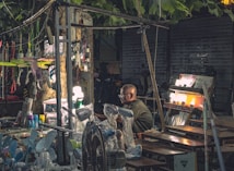 A market stall located outdoors under the shade of trees, displaying various electrical appliances and goods. The stand includes items like fans and lamps, with some being illuminated. The vendor, an elderly individual wearing glasses, sits amidst the array of products. The scene is dimly lit, with artificial lighting enhancing the visibility of the merchandise. The background is urban, featuring metallic shutters and parked bicycles.