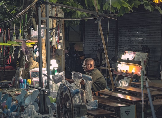 A market stall located outdoors under the shade of trees, displaying various electrical appliances and goods. The stand includes items like fans and lamps, with some being illuminated. The vendor, an elderly individual wearing glasses, sits amidst the array of products. The scene is dimly lit, with artificial lighting enhancing the visibility of the merchandise. The background is urban, featuring metallic shutters and parked bicycles.