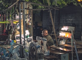 A market stall located outdoors under the shade of trees, displaying various electrical appliances and goods. The stand includes items like fans and lamps, with some being illuminated. The vendor, an elderly individual wearing glasses, sits amidst the array of products. The scene is dimly lit, with artificial lighting enhancing the visibility of the merchandise. The background is urban, featuring metallic shutters and parked bicycles.