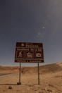 A brown road sign stands in a desert landscape under a clear blue sky. The sign reads 'Ruta del Desierto, Cordillera De La Sal, Desierto y Arqueología' and features icons depicting hiking, landscape, and photography. The barren, rocky terrain stretches into the distance with a road curving away.