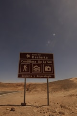 A brown road sign stands in a desert landscape under a clear blue sky. The sign reads 'Ruta del Desierto, Cordillera De La Sal, Desierto y Arqueología' and features icons depicting hiking, landscape, and photography. The barren, rocky terrain stretches into the distance with a road curving away.