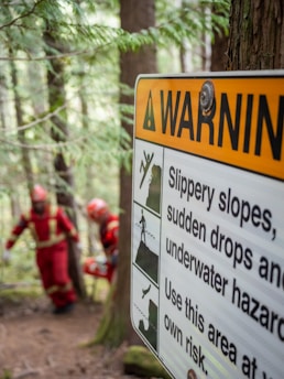 A warning sign posted on a tree in a forested area. The sign includes text and symbols cautioning about slippery slopes, sudden drops, and underwater hazards. In the background, two people wearing red protective suits are walking along a forest path.