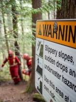 A warning sign posted on a tree in a forested area. The sign includes text and symbols cautioning about slippery slopes, sudden drops, and underwater hazards. In the background, two people wearing red protective suits are walking along a forest path.