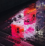 A colorful still from a music video showing vibrant city lights reflecting on wet pavement.