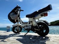 A scooter is parked on a concrete surface near a large body of water. The sky is clear with a few clouds, suggesting a sunny day. The scooter is white with a black seat and has a basket attached to the front. In the background, trees and a blue ocean are visible.