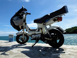 A scooter is parked on a concrete surface near a large body of water. The sky is clear with a few clouds, suggesting a sunny day. The scooter is white with a black seat and has a basket attached to the front. In the background, trees and a blue ocean are visible.