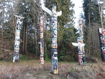A collection of intricately carved and painted totem poles surrounded by trees. The totem poles feature vibrant colors including blue, red, and green, depicting various figures and symbols. The surrounding area is a forest with tall, dense trees and undergrowth.