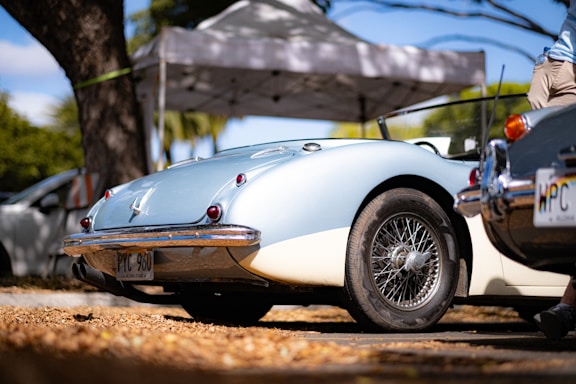 A vintage convertible car with a light blue and white exterior is parked under a canopy. The license plate reads 'PYC 960'. The car features classic wire-spoke wheels and red tail lights. There is another car partially visible in the foreground, along with a person standing next to it.