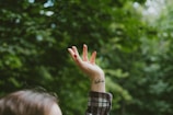 A hand with a tattoo reaches upward, framed against a blurred green forest background. The fingers are gently splayed and the person is wearing a plaid shirt. Light filters through the leaves, creating a serene atmosphere.