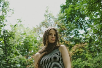 A person with long hair stands outdoors in a natural setting, surrounded by lush green trees and foliage. They are wearing a striped sleeveless top, and the lighting is soft, suggesting a serene, peaceful atmosphere.