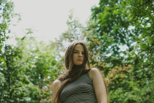 A person with long hair stands outdoors in a natural setting, surrounded by lush green trees and foliage. They are wearing a striped sleeveless top, and the lighting is soft, suggesting a serene, peaceful atmosphere.
