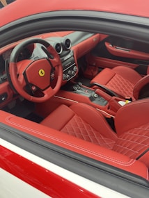The image features a luxury car interior with red leather upholstery and black accents. The dashboard is sleek and modern, with a prominent logo in the center of the steering wheel. The seats are quilted for added luxury, and there are several buttons and controls within easy reach.