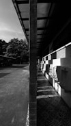 A black and white image of an outdoor facility with urinals lined up against a wall under an overhanging roof. The scene is split symmetrically by a vertical pillar, with sunlight casting strong shadows on the patterned floor.