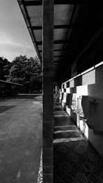 A black and white image of an outdoor facility with urinals lined up against a wall under an overhanging roof. The scene is split symmetrically by a vertical pillar, with sunlight casting strong shadows on the patterned floor.